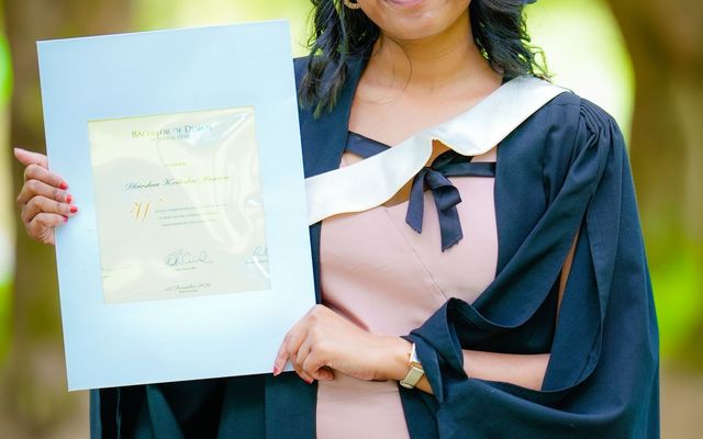 A graduate in cap and gown holding up her certificate outdoors