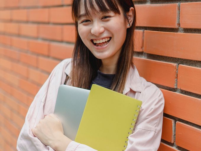 A smiling student holding notebooks by a brick wall