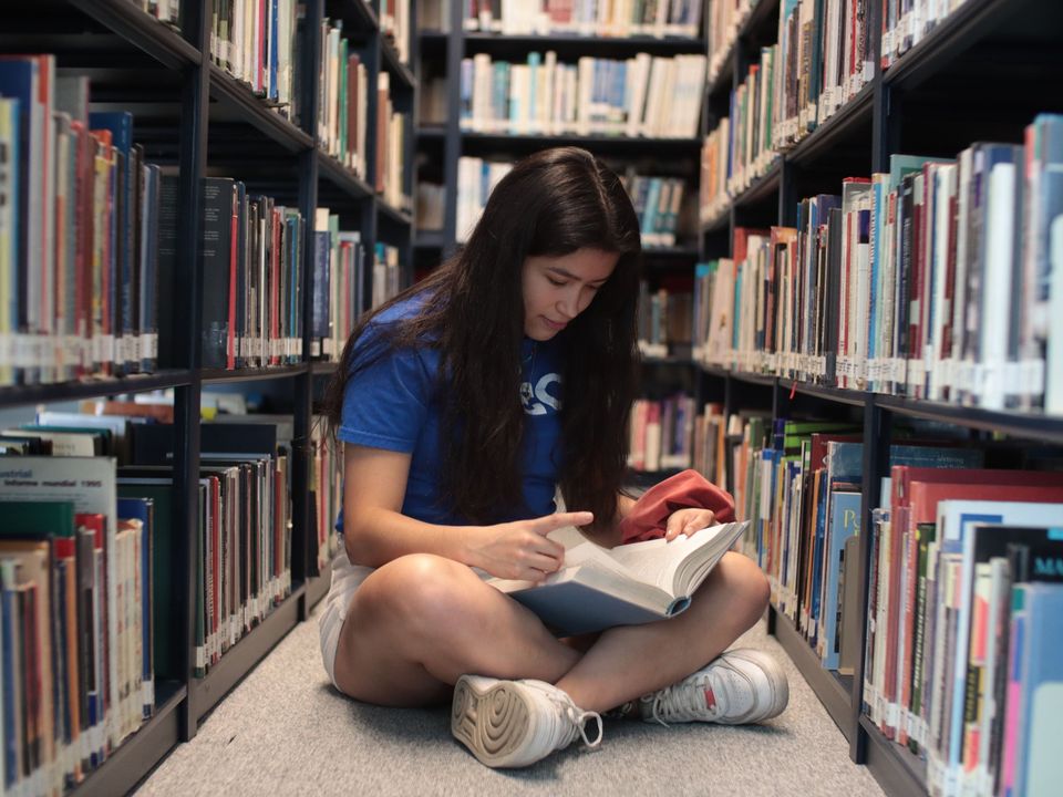 A student sitting on a library floor, reading a book between the shelves