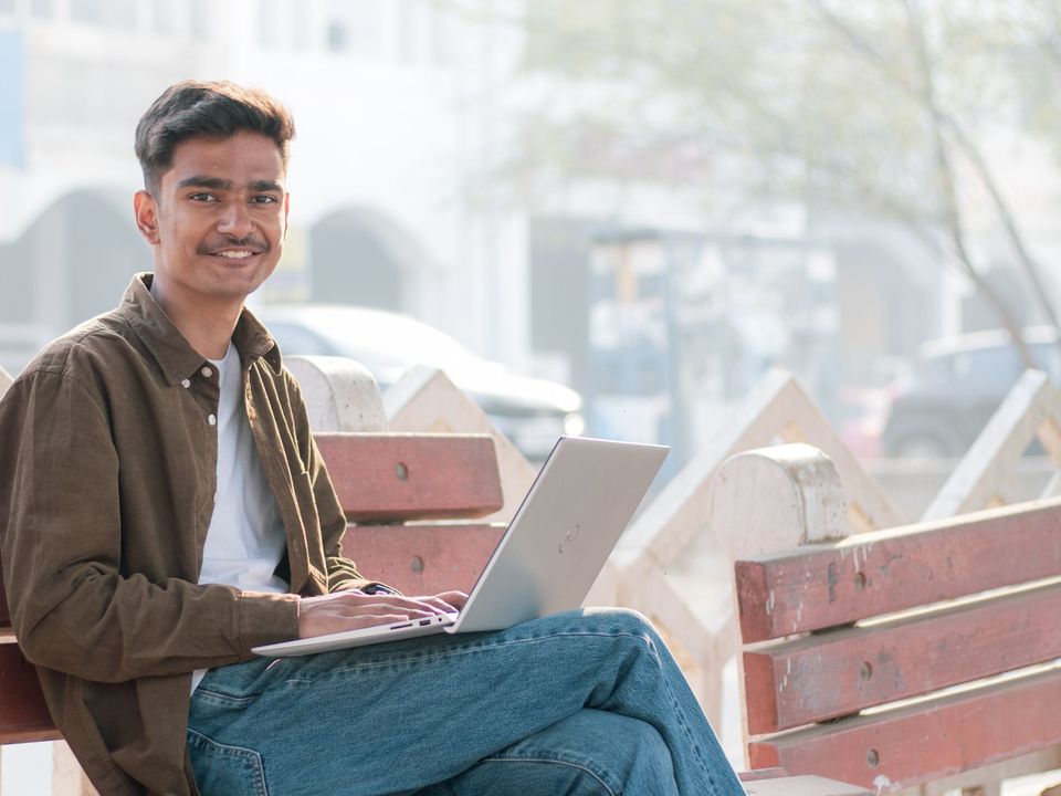 Young man on a bench outdoors using a laptop, smiling at the camera.